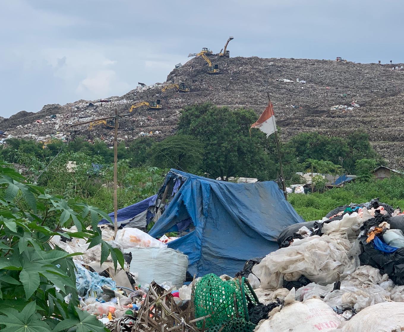 A shack carring the Indonesian flag nestles in greenery beneath the landfill.