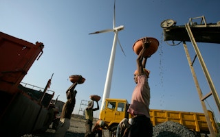 A laborer hauls crushed rocks pulversied by a diesel-powered crusher in the shadow of a wind turbine.