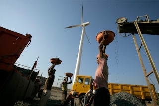 A laborer hauls crushed rocks pulversied by a diesel-powered crusher in the shadow of a wind turbine.